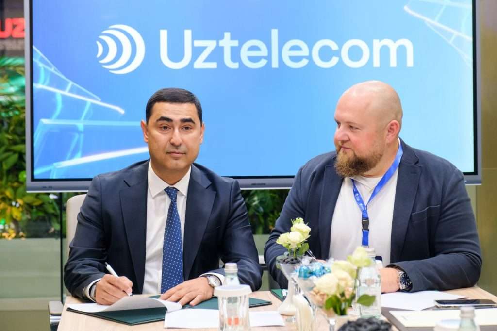 A business meeting with two men seated at a table in front of a large Uztelecom display. Both are wearing conference badges and reviewing documents.
